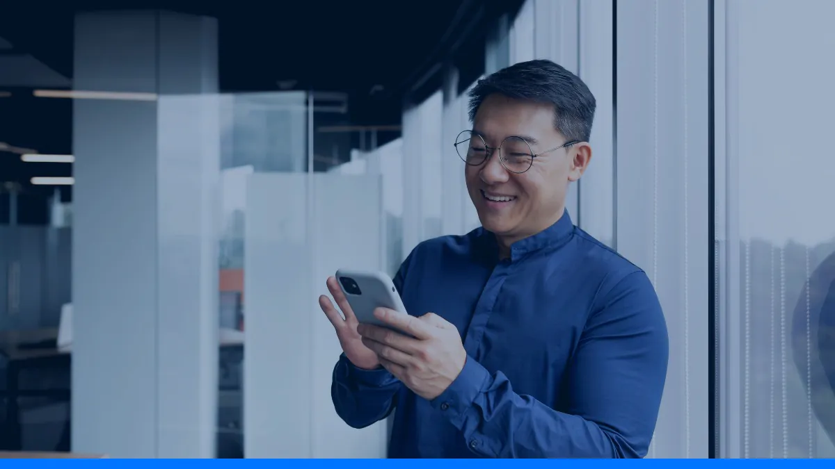 Homem sorrindo enquanto lê o celular perto da janela de um escritório moderno